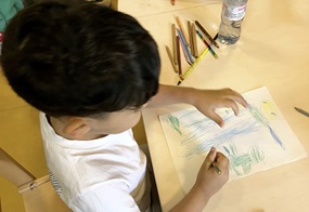 Ein Kind zeichnet mit Buntstiften ein Bild auf ein Papier.