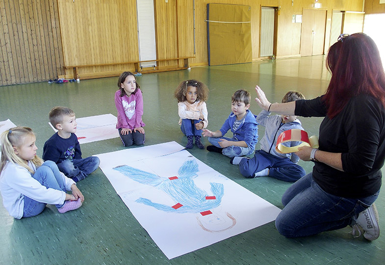Kinder sitzen am Boden und schauen sich eine große Zeichnung von einem Körper an