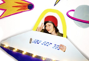 Aus einem Ufo aus Pappe schaut eine Frau mit einem roten Helm heraus.