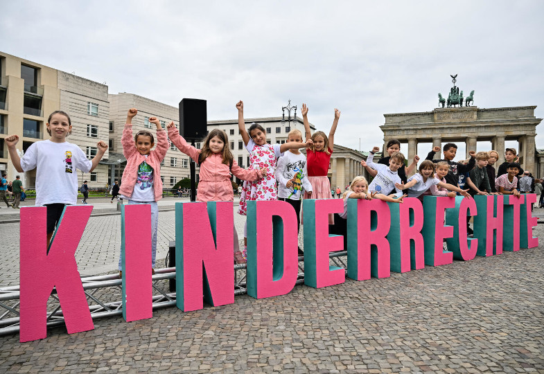 Mehrere Kinder stehen hinter einem großen Kinderrechte-Logo.