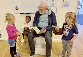 Eine erwachsene Person sitzt mit einem Buch in der Hand auf einem Stuhl und liest daraus vor, während drei Kinder lachend daneben stehen oder sitzen.