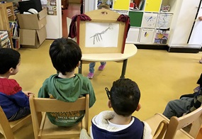 Um ein Kind, das ein gemaltes Bild zeigt, sitzen viele Kinder auf Stühlen.