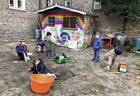 Mehrere Kinder graben auf einem Hinterhof in einem mit Absperrband markierten Bereich, während im Hintergrund ein bunt bemaltes Gartenhäuschen zu sehen ist.