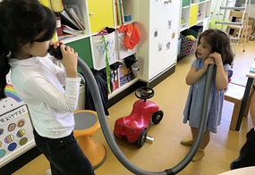 Zwei Kinder halten sich ein ausrangiertes Staubsaugerrohr ans Ohr. 