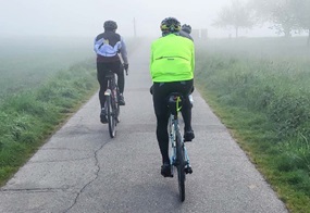 Drei Menschen fahren Fahrrad auf einem Radweg, der durch neblige Wiesen führt