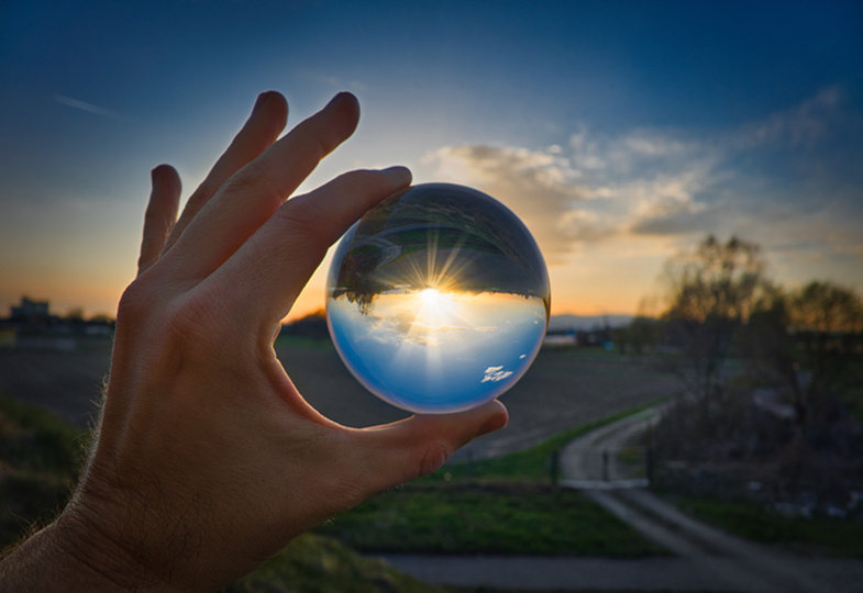 im Hintergrund geht die Sonne unter, eine Hand halt eine Glaskugel zwischen zwei Fingern, in der sich die Sonnenstrahlen brechen