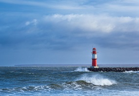 Ein roter Leuchtturm steht auf einer Steinmole am stürmischen Meer, während Wellen gegen die Küste schlagen.