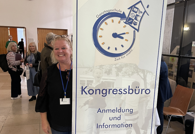 Eine Frau steht hinter einem großen Schild, auf welchem "Ganztagsschule Kongressbüro" steht.