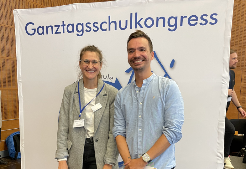 Ein Mann und eine Frau vor einem großen Schild mit der Aufschrift "Ganztagsschilkongress".