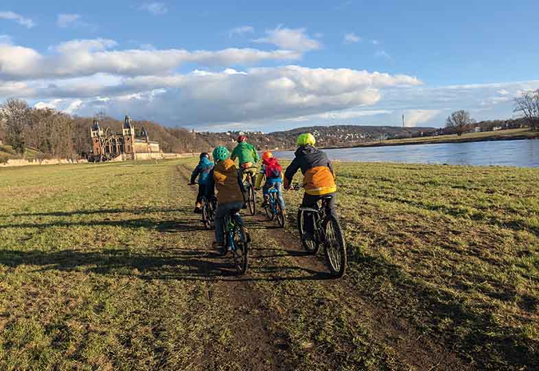 Fünf Personen, eine Mischung aus Kindern und Erwachsenen, fahren auf ihren Fahrrädern entlang eines Feldwegs, der sich parallel zu einem Fluss erstreckt. 