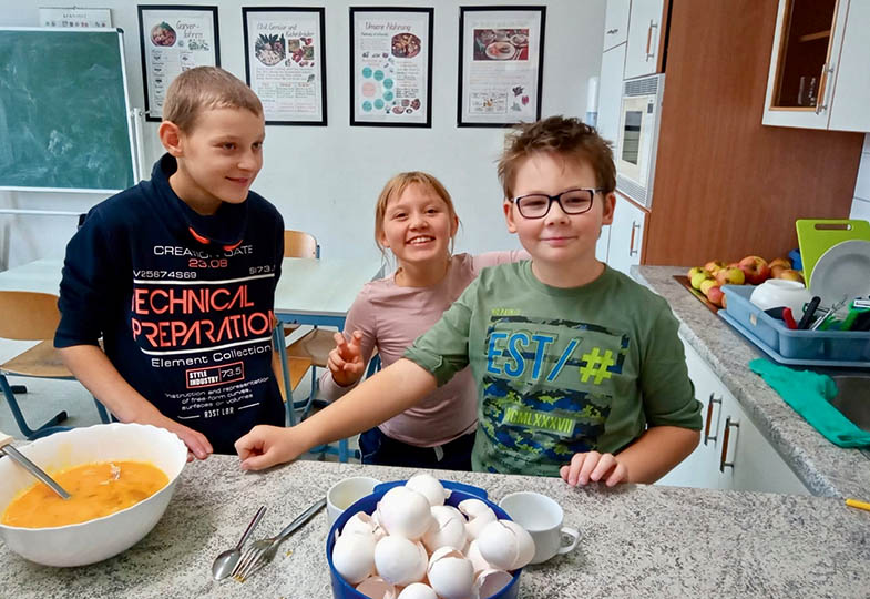 Ein Mädchen und zwei Jungen stehen in einer Schulküche. Vor ihnen stehen eine Schüssel mit Eierschalen und eine weitere Schüssel mit verquirltem Ei.