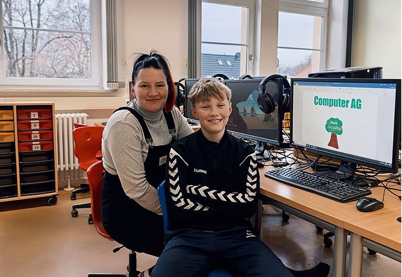 Ein Junge und eine Frau sitzen in einem Computerraum und schauen lächelnd in die Kamera.