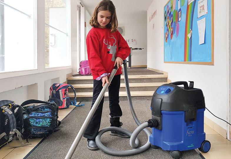 Ein Mädchen hält einen Staubsauger in der Hand und steht im Flur einer Schule. Auf dem Boden liegen mehrere Rucksäcke.