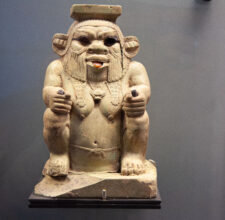 Foto einer altägyptischen Bes-Vase aus Ton, bauchig geformt, mit plastischem Gesicht des Gottes Bes: breite Nase, große Augen, Löwenmähne und herausgestreckte Zunge