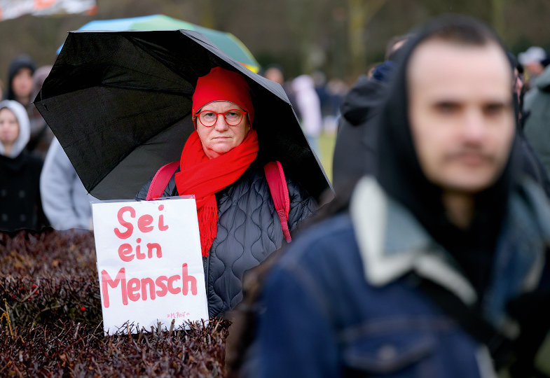 Eine grimmig guckende Demonstrantin hält einen Regenschirm und ein Schild mit der Aufschrift "Sei ein Mensch"