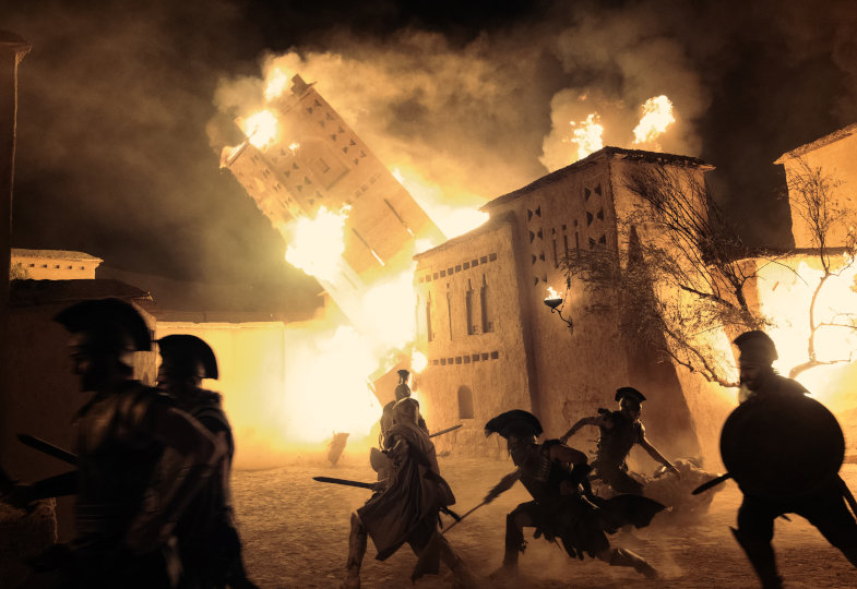 Kampfszene bei Nacht. Soldaten kämpfen im Vordergrund, während im Hintergrund ein brennender Turm einstürzt. 