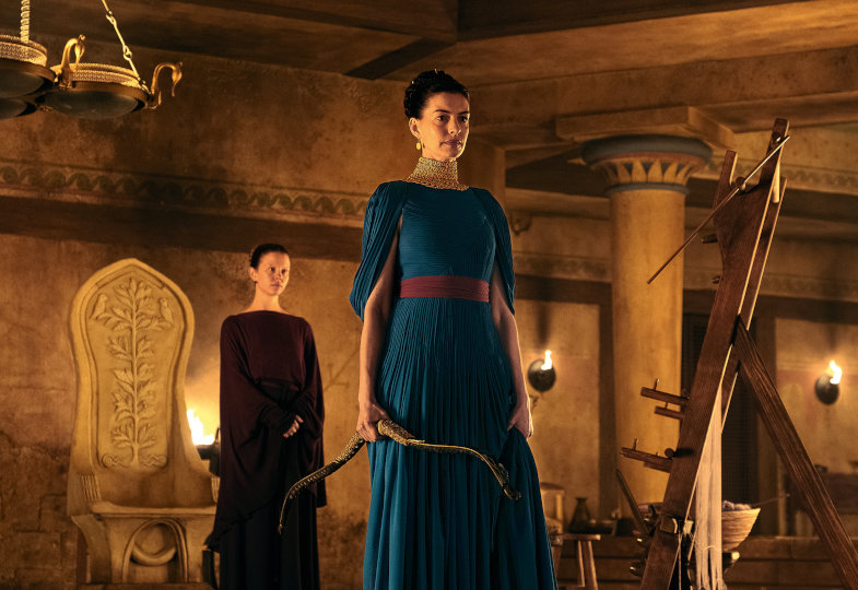 Eine braunhaarige, junge Frau mit einem blauen Kleid hält einen Bogen in der Hand