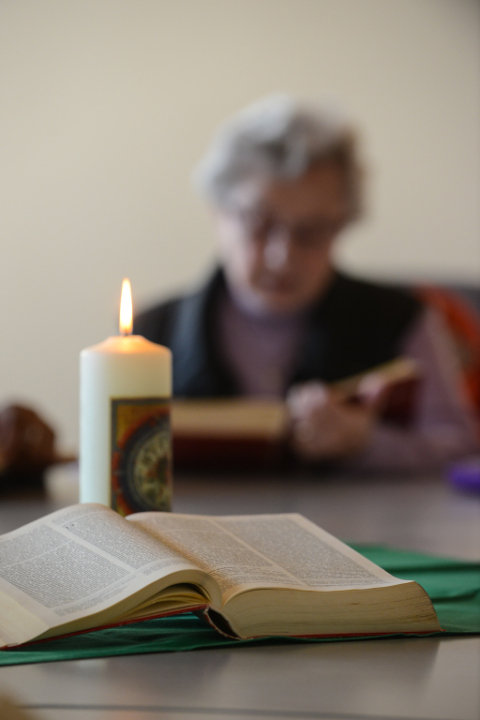 Eine aufgeschlagene Bibel und eine brennende Kerze auf einem Tisch, im Hintergrund liest eine Frau in einer Bibel