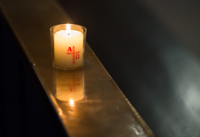 Eine brennende Kerze mit Kreuz und Alpha-Omega-Symbolen steht auf einer Kirchenbank