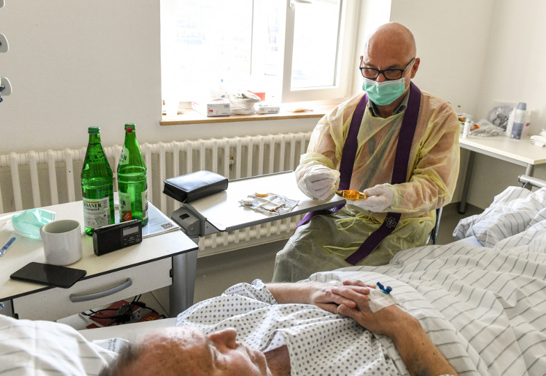 Ein Priester in klinischer Schutzkleidung spendet einem Patienten die Krankenkommunion