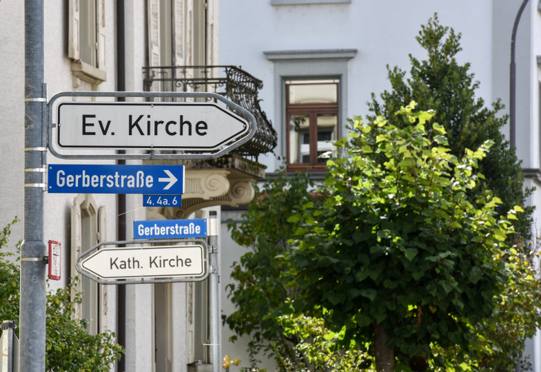 Zwei Straßenschilder weisen - in entgegengesetzter Richtung - den Weg zu einer evangelischen und einer katholischen Kirche.