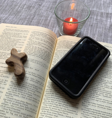 Aufgeschlagene Bibel, darauf ein Kreuz und ein Smartphone, daneben eine brennende Kerze