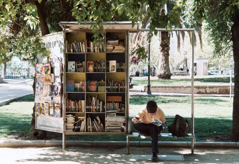Ein junger Mensch sitzt umgeben von einem Bücherregal lesend an einer Haltestelle