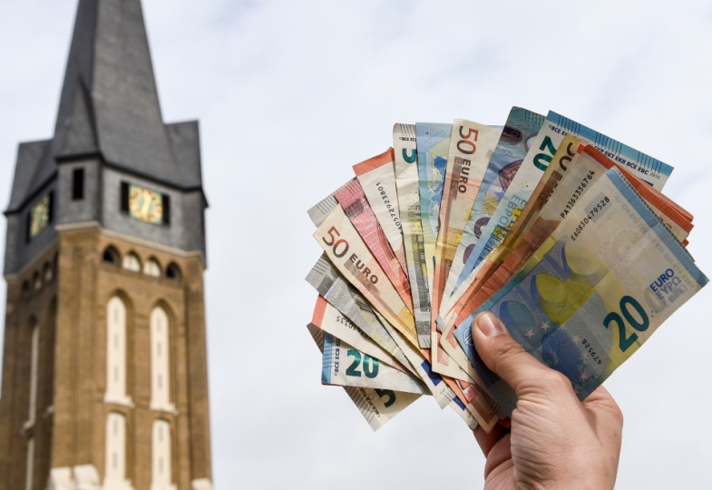 Eine Hand hält ein Bündel Geldscheine neben einem Kirchturm in die Luft.
