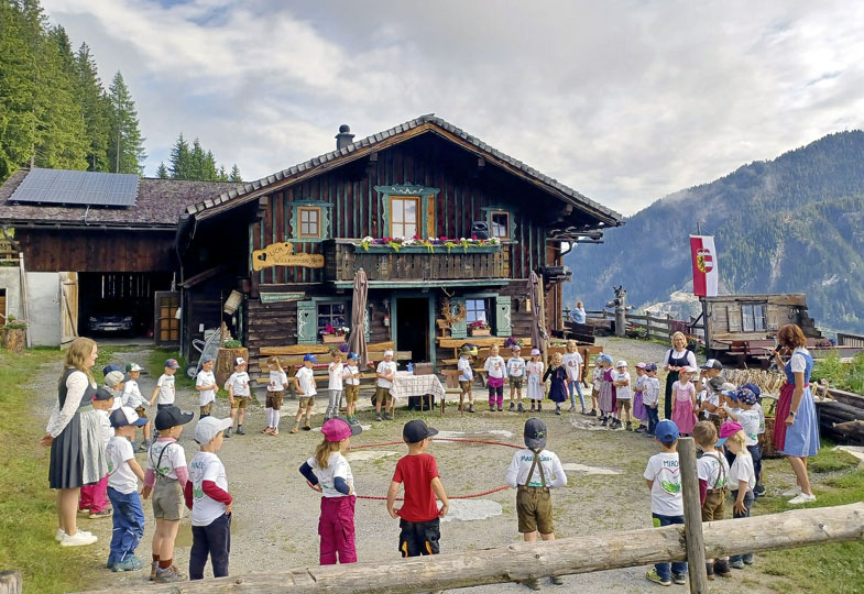 Im Hintergrund ein Holzhaus (die Alm) mit Bäumen und einem Berg, davor stehen Kinder und Erwachsene in einem Kreis auf dem Hof.