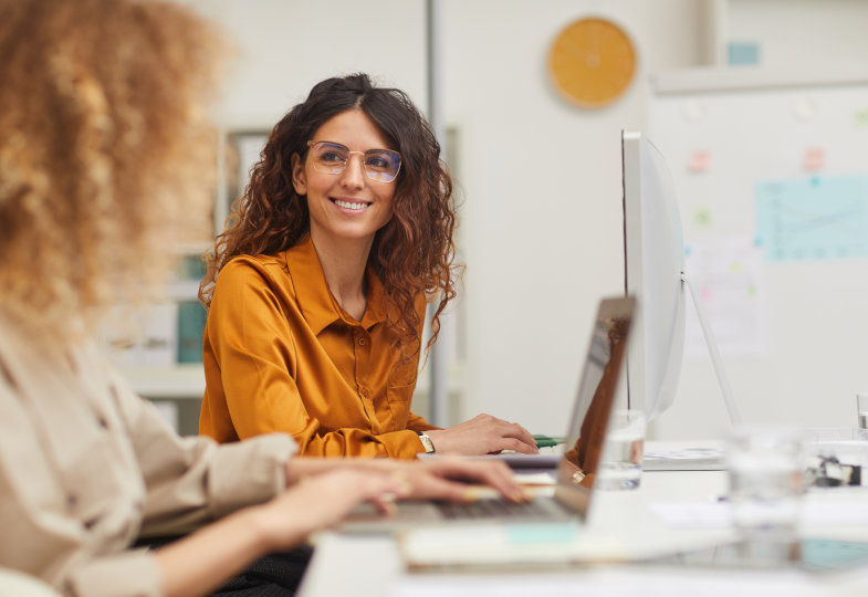 Zwei Frauen sitzen an einem Schreibtisch mit Computern.