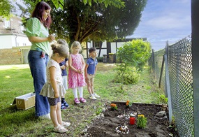 Vier Kinder und eine Erzieherin stehen im Garten vor einem Grab, das Blumen und eine Kerze enthält.