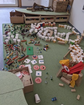 Eine auf einem grünen Teppich aufgebaute, abgegrenzte Miniaturlandschaft aus Alltagsgegenständen wie Pappbechern, Pappdeckeln, Spielzeugtieren und Spielzeugautos.
