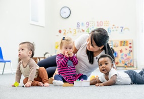 Drei Kleinkinder sitzen auf dem Boden, hinter Ihnen ist eine Fachkraft.