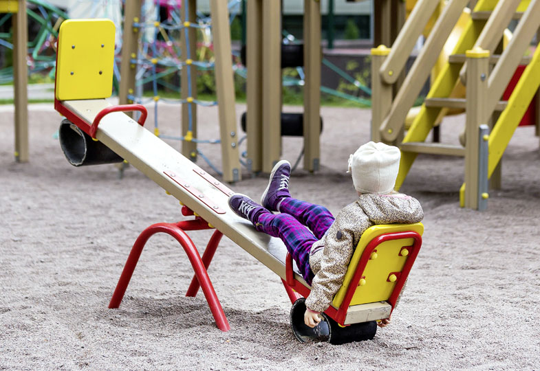 Ein Spielplatz mit einer Wippe im Vordergrund, auf einer Seite sitzt ein Kind mit dem Rücken zur Kamera.