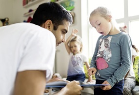 Ein Mädchen liest gemeinsam mit einer erwachsenen Person aus einem Buch, während sich im Hintergrund ein weiteres Kind befindet.
