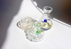 Drei unterschiedlich geformte Glasgefäße, gefüllt mit Flüssigkeit, in denen jeweils eine farbige Murmel liegt.