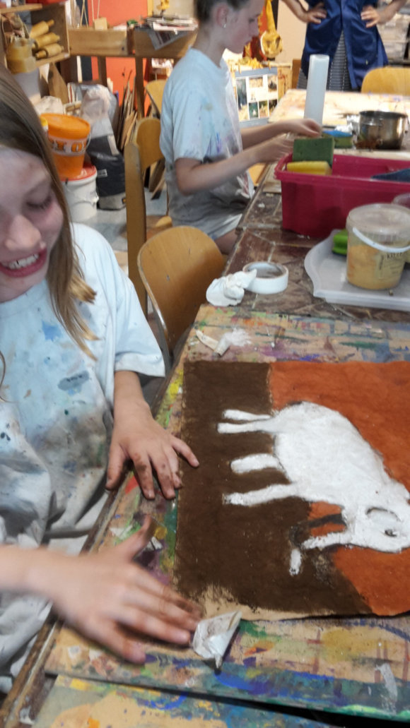 Ein Mädchen sitzt vor einem Gemälde in Steinzeit-Optik, dass einen Mammut darstellt.