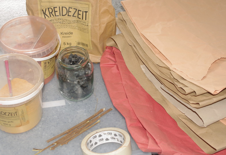 Rechts liegt braunes Papier, links Kunststoffbehälter mit braunem Pulver, davor Stöcke, Kreppband und ein Glas mit schwarzen Steinen.