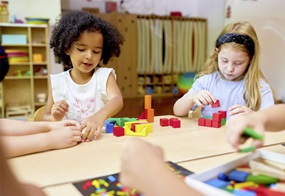 Zwei Kinder sind am Tisch ins Konstruieren vertieft und spielen mit bunten Bauklötzen.