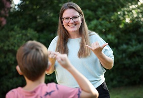 Eine Frau lächelt und spricht in Gebärdensprache mit einem Kind, das nur von hinten zu sehen ist.