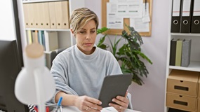 Eine Frau sitzt in einem Büro und schaut konzentriert auf ihr Tablet, welches sie in der Hand hält.