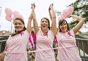 Drei Frauen mit einer rosa Schleife und rosa T-Shirts heben freudig die Arme nach oben.