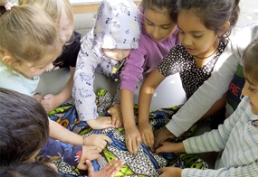 Viele Kinder sitzen im Kreis, in dessen Mitte eine Decke einen Gegenstand verdeckt, den sie ertasten sollen.