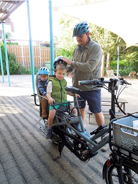 Zwei Kinder sitzen auf einem stehenden Lastenrad, und ein Erwachsener setzt einem von ihnen einen Helm auf.