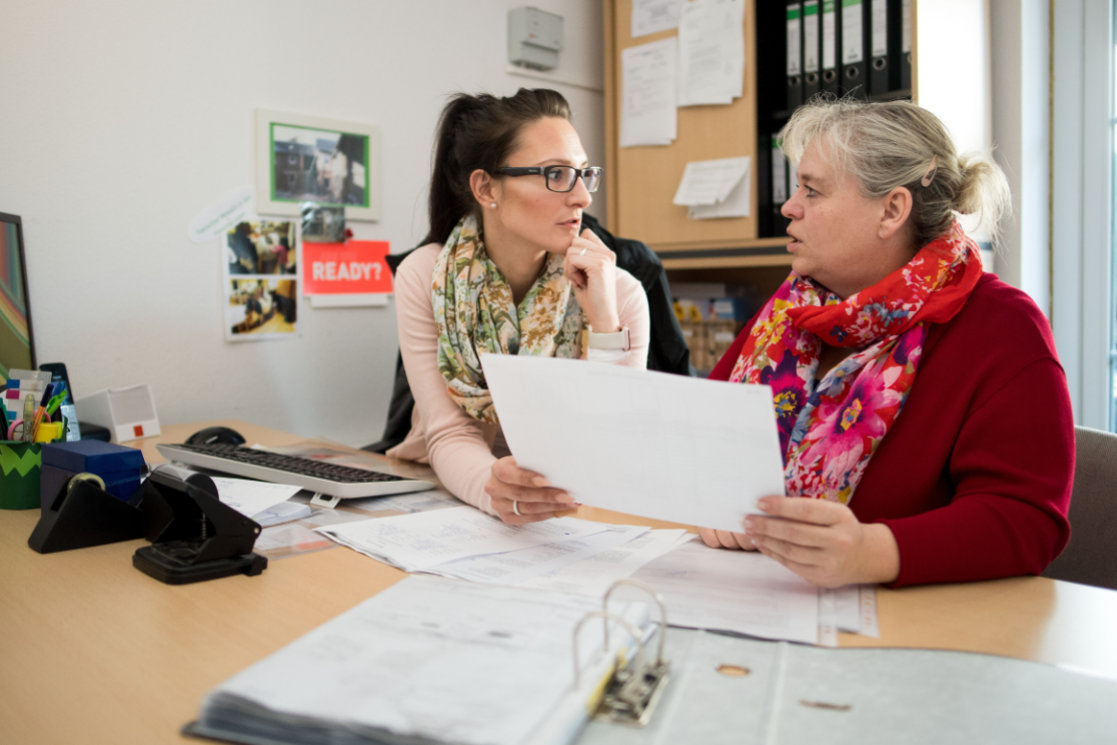 Zwei Frauen sitzen gemeinsam an einem Tisch in einem Büro, halten ein Blatt Papier in den Händen und wirken in ein angeregtes Gespräch darüber vertieft.