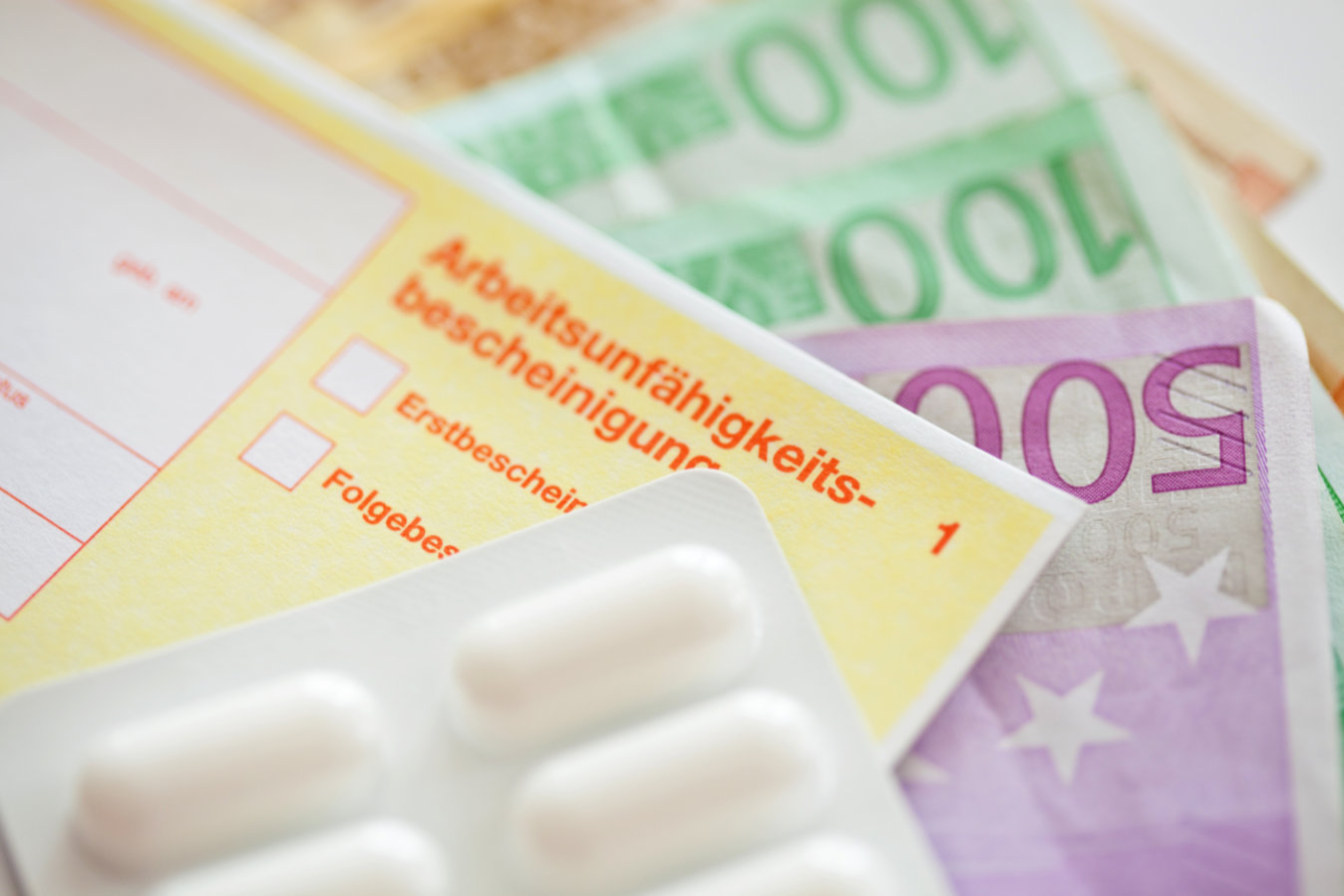 Mehrere Geldscheine liegen unter einer Arbeitsunfähigkeitsbescheinigung, auf der ein Tabletten-Blister platziert ist.