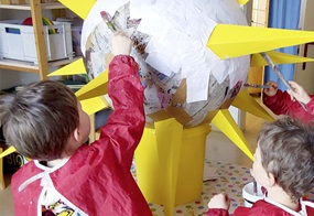 Kinder kleistern weißes Papier an eine Kugel, die mit gelben Pappspitzen eine Sonne darstellen soll.