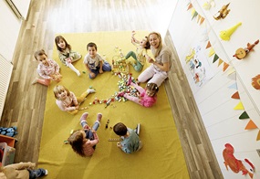 Die Kinder sitzen auf einem gelben Teppich und spielen mit verschiedenen Bauklötzen um sich herum.