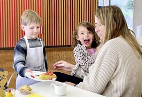 Ein Kind mit Schürze bringt einen Teller mit Erdbeerkuchen an den Tisch, an dem bereits eine Frau und ein weiteres Kind sitzen.