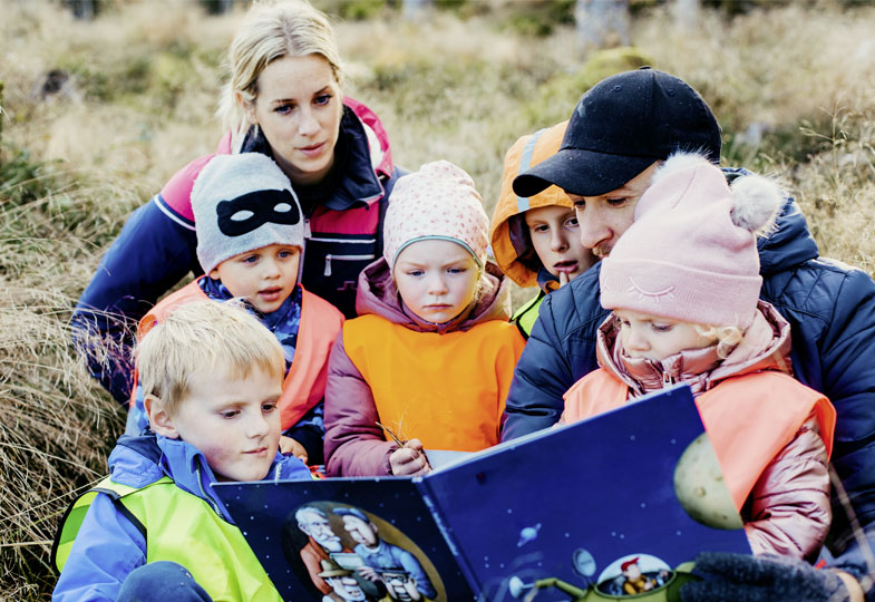 Kinder und Erwachsene sitzen gemeinsam auf einer Wiese, schauen zusammen in ein Buch und lesen miteinander.